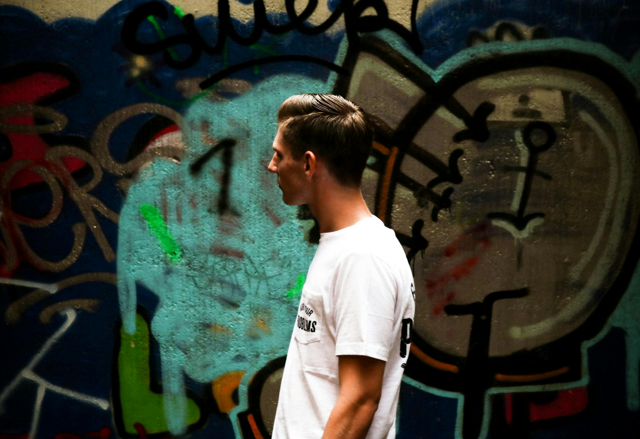 Side profile of a young man in a white t-shirt walking past a colorful graffiti wall.